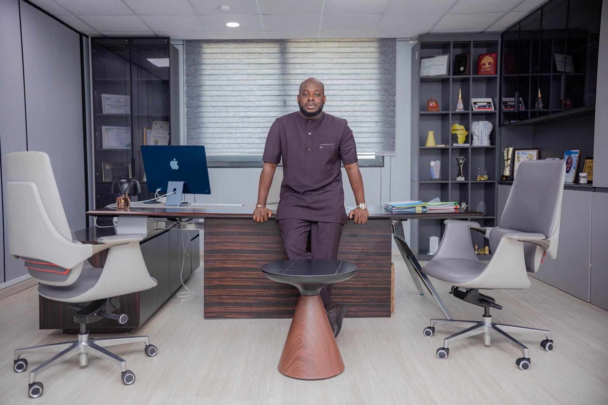 Bakary DIABY debout dans son bureau de direction, appuyé à son grand bureau, entre ses certificats et ses trophées